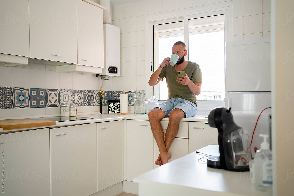 Man drinking a coffee cup and using his phone at kitchen