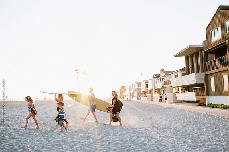 Family walking on sand in beach holding surfboards and bags and skateboards at golden hour