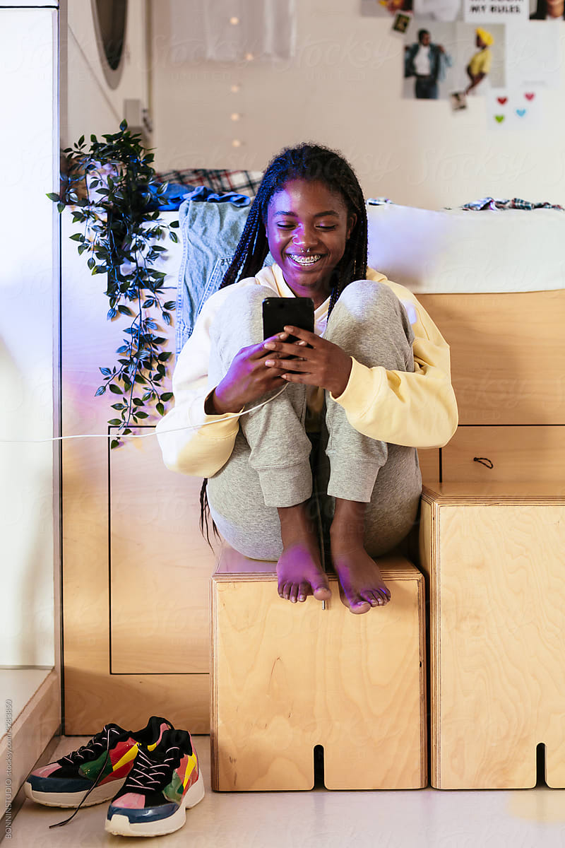 Happy black teenager using smartphone near bed