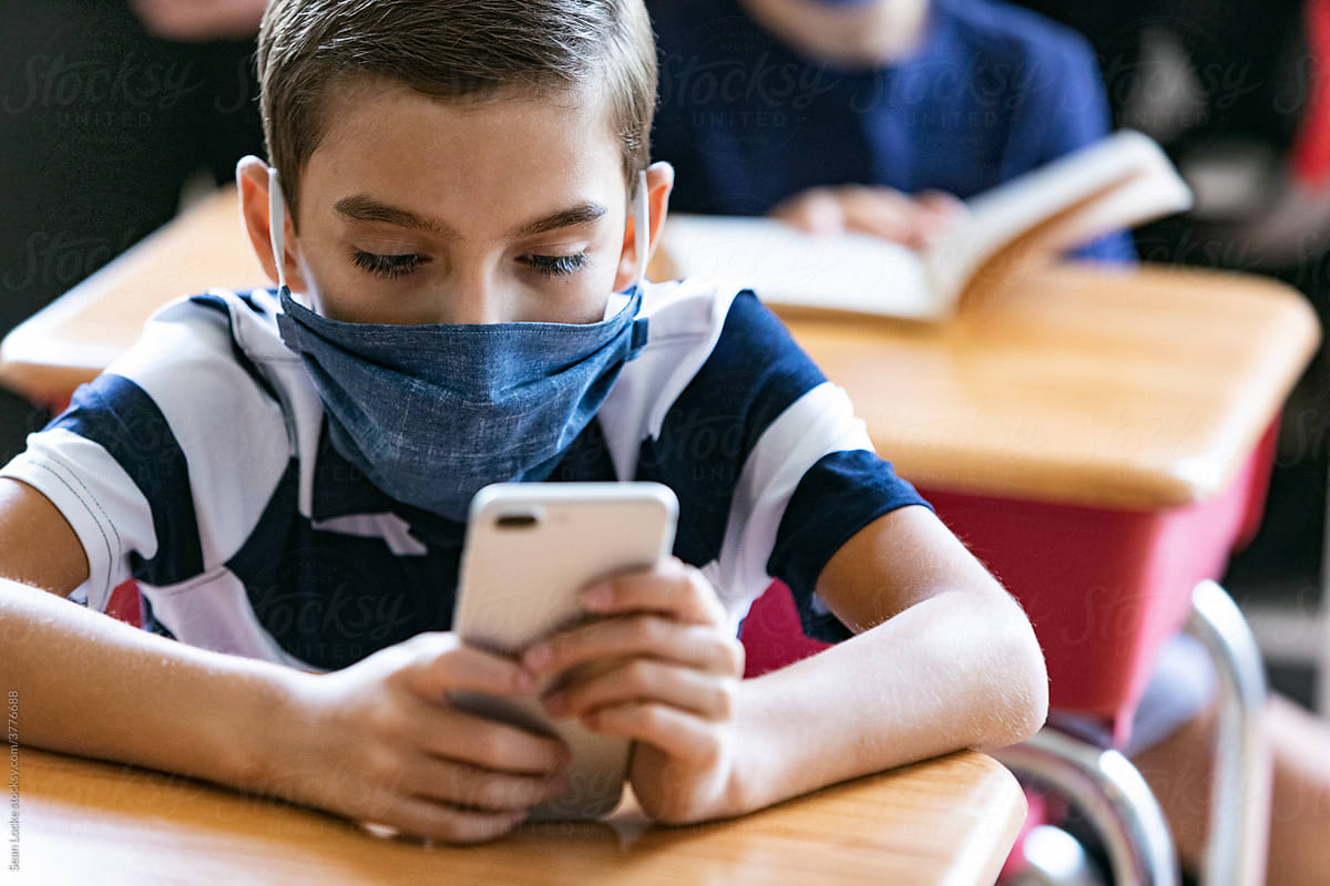 School: Student Wearing Face Mask Checks Cell Phone At Desk