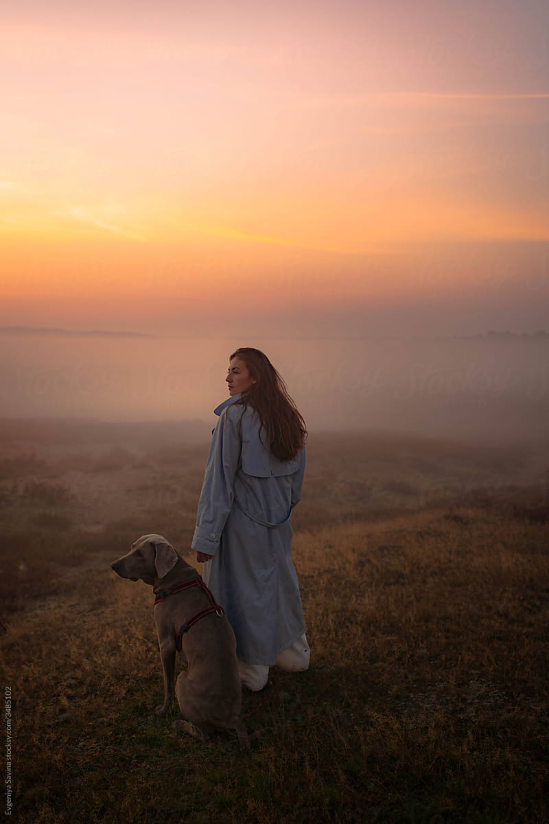 A girl with a long hair standing in the middle of the foggy and misty field with a dog in the early morning