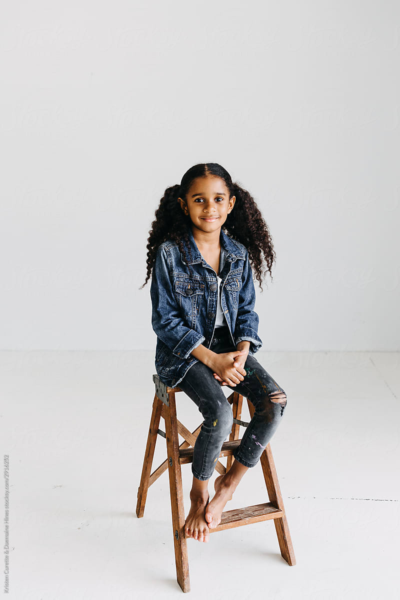 Studio portrait of a black girl sitting on a step stool with white background.