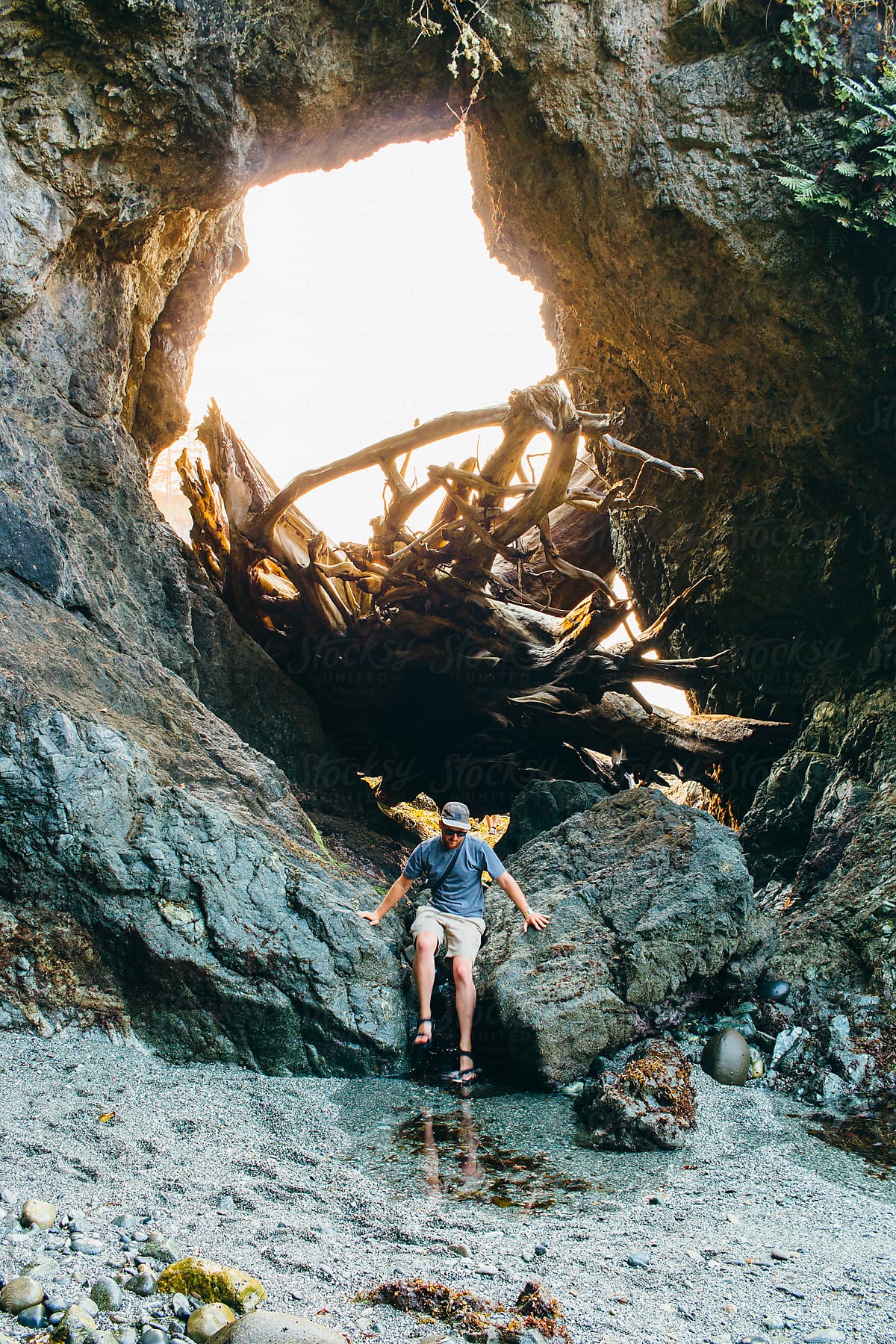 Young Man Wearing Shorts And Sandals Climbing Through Rocky Arch On Ocean shore