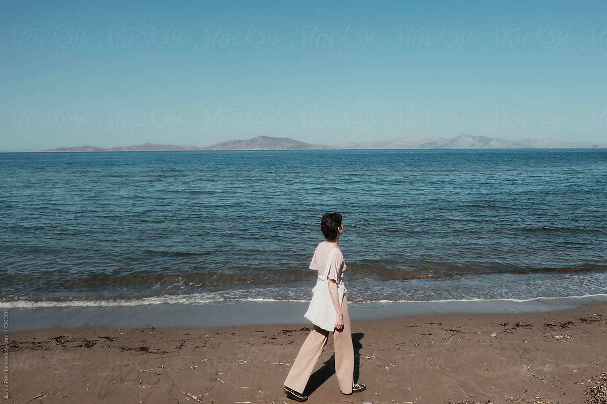 A woman with a short haircut and a bag walks on the sand.
