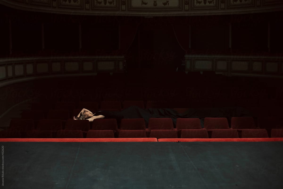 Young woman in a long black dress lies down on a theater chairs