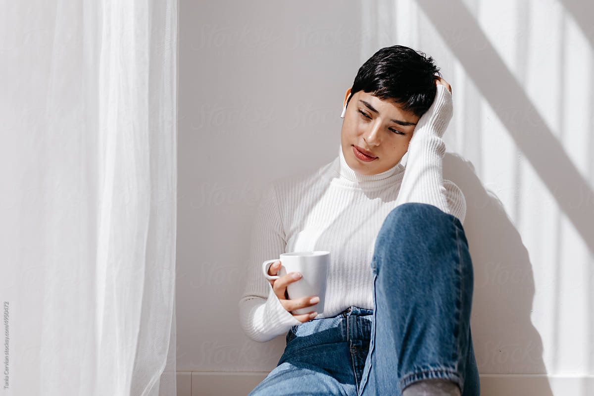 Pensive lady in TWS earphones drinking coffee on floor