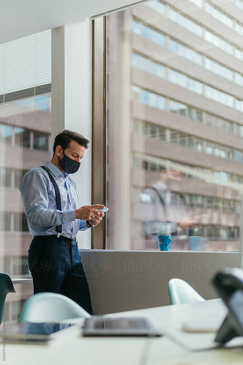 Serious office worker in mask using smartphone at workplace