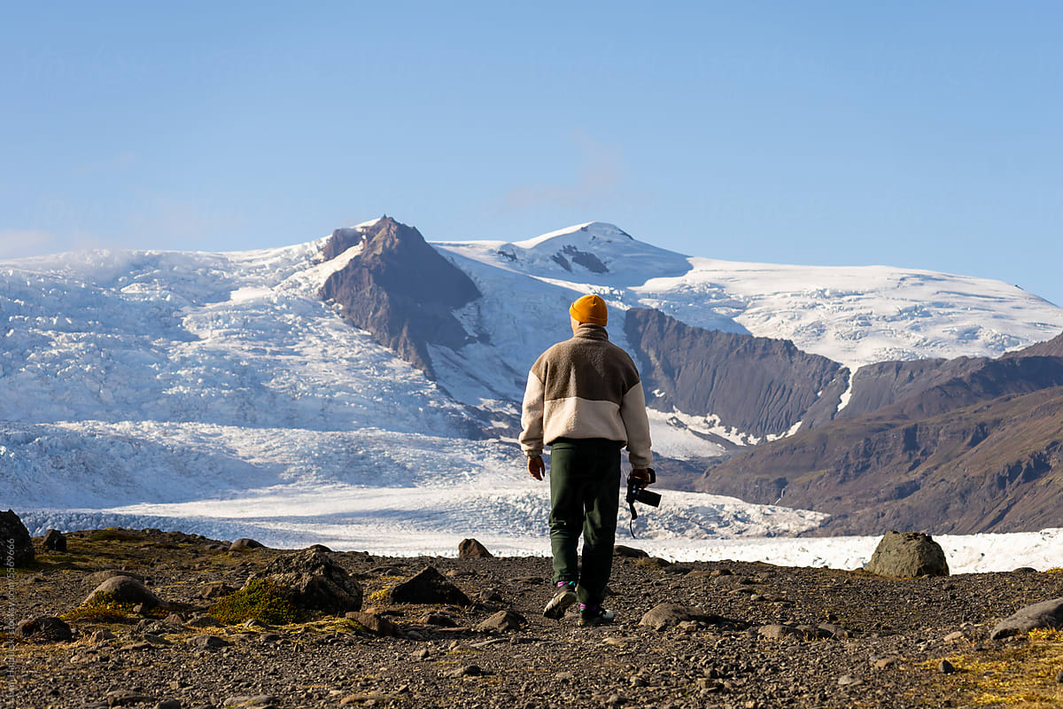 young man from behind holding camera in hands walking to huge glacier