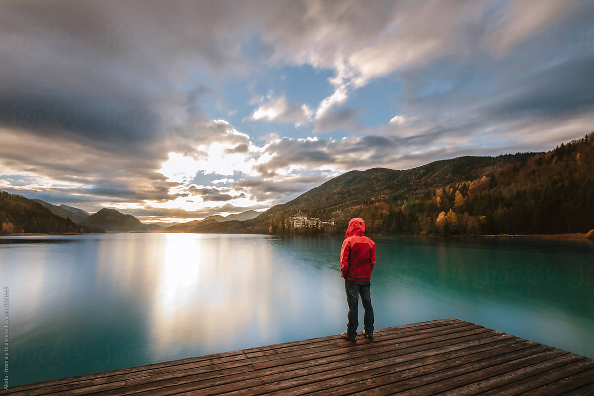 young man in red jacket standing on a jetty  at a lake enjoying the sunrise