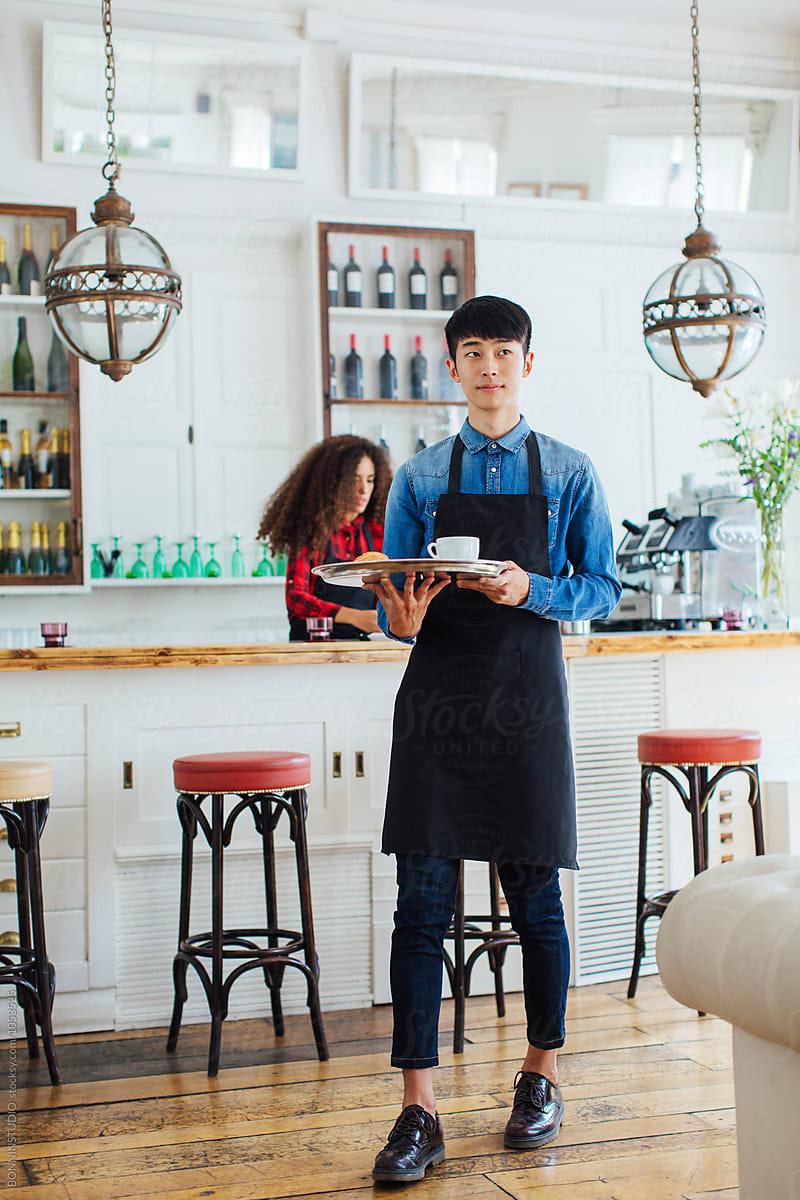 Young asian man wearing an apron holding a tray with coffee in a beautiful coffee shop.