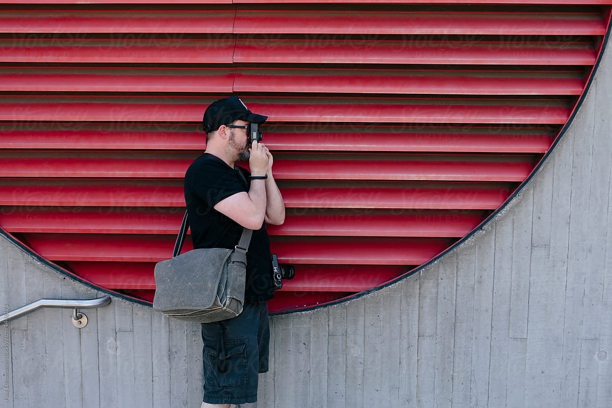Street photographer taking photos with film camera, red industrial wall behind
