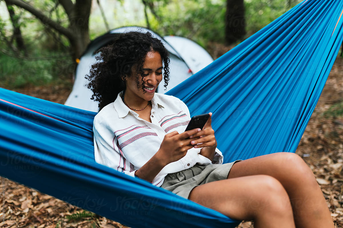 Cheerful curly hair woman smiling and laughing while she is using her smartphone, seated on a blue hammock