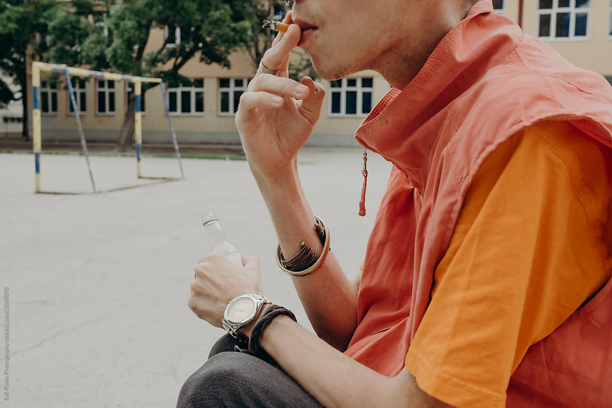 Anonymous young skinny male model drinking alcohol and smoking cigarette at the school yard