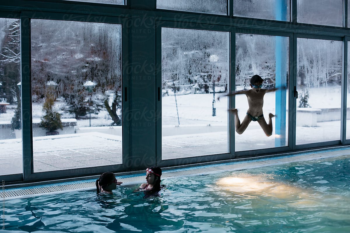 Boy jumping into the indoors swimming pool with lots of windows with view of a winter landscape