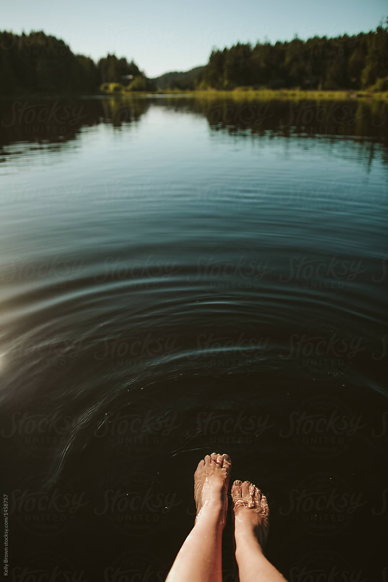 a young woman\'s feet dangle into the water of a lake