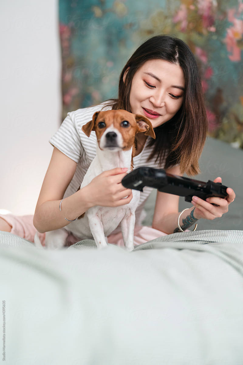 Woman playing videogame on bed with dog