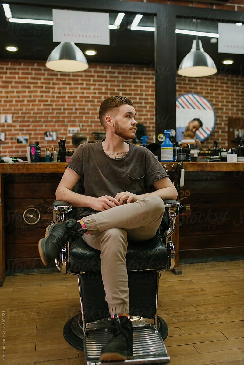 A ginger haired guy sits on the chair in the barbershop