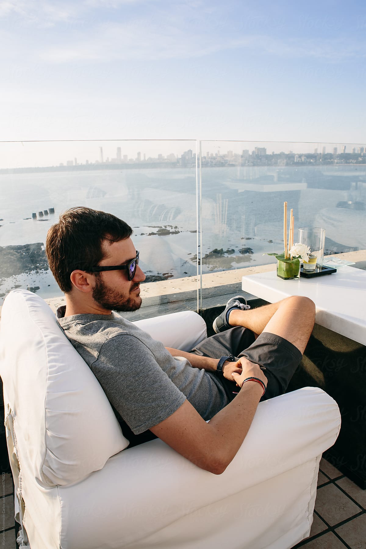 Young man sitting down on a sofa on a lounge cafe bar on the rooftop of a city at sunset