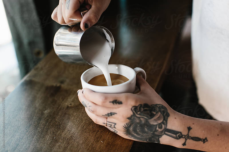 hands with tattoos pouring latte art in white mug