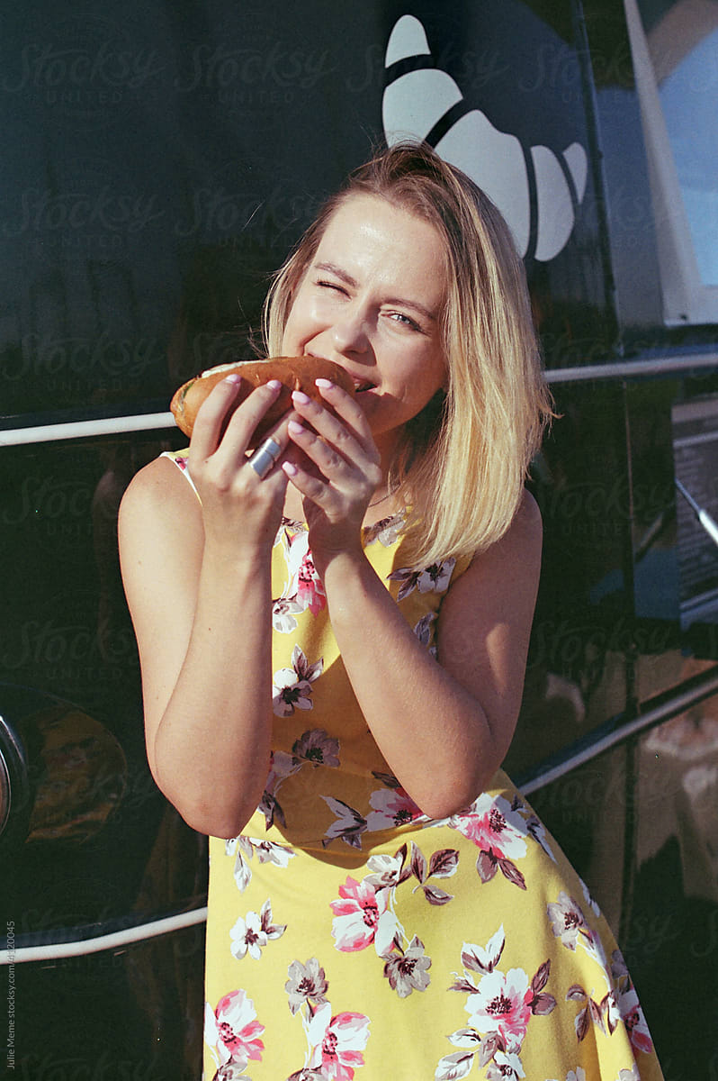 Blond girl eats hot-dog on a sunny day near the van