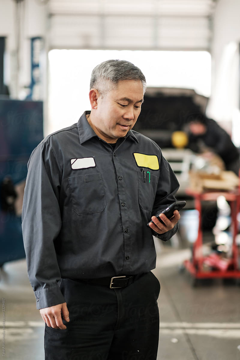 Asian car mechanic checking his phone in his auto repair shop