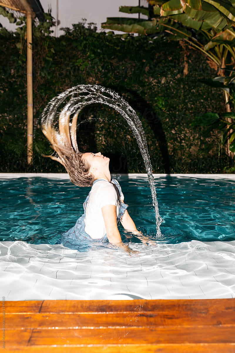Young woman flicks hair with water in the air while wearing all her clothes in the pool
