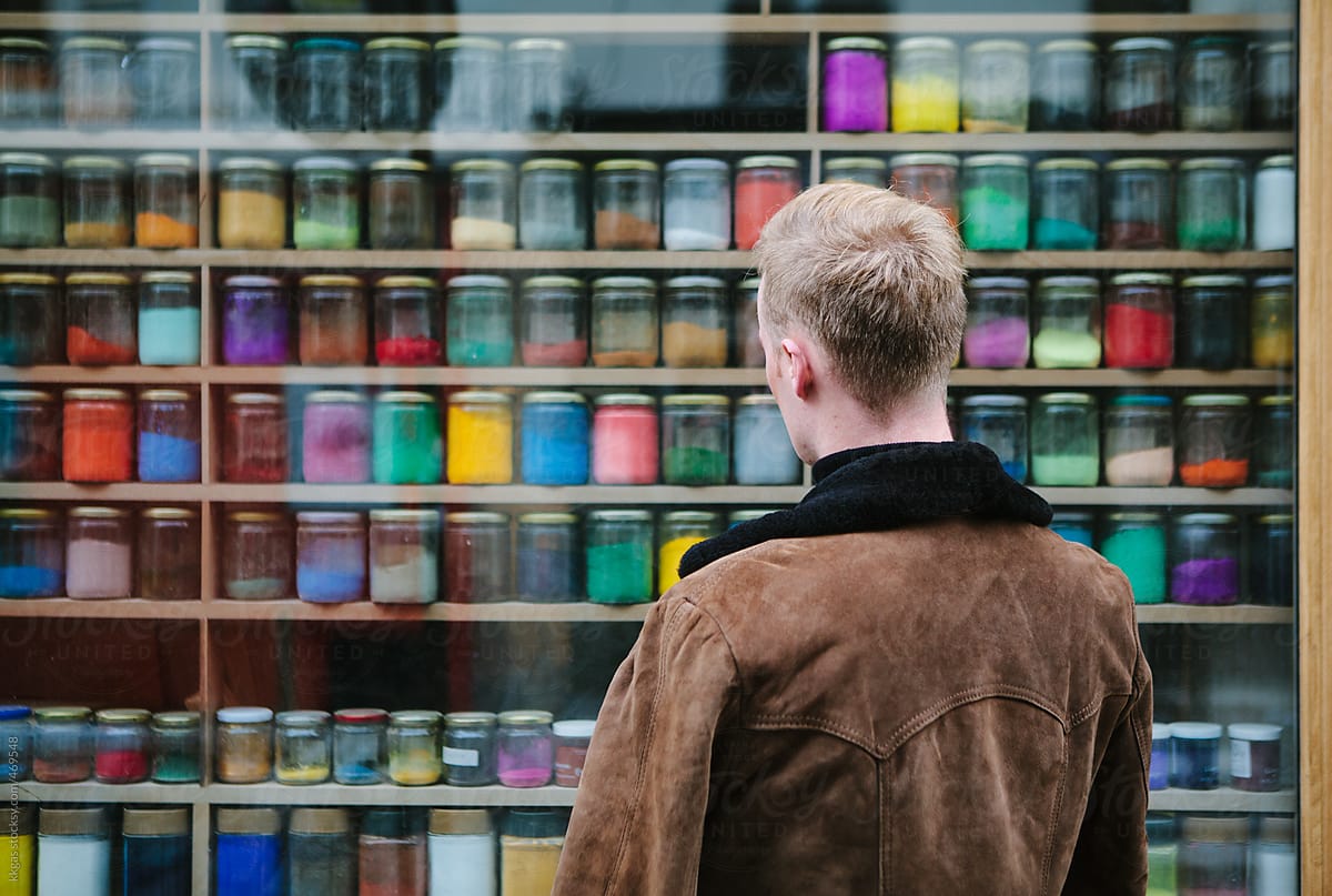 Blonde man next to a shop window filled with artists powder paints of many colours.