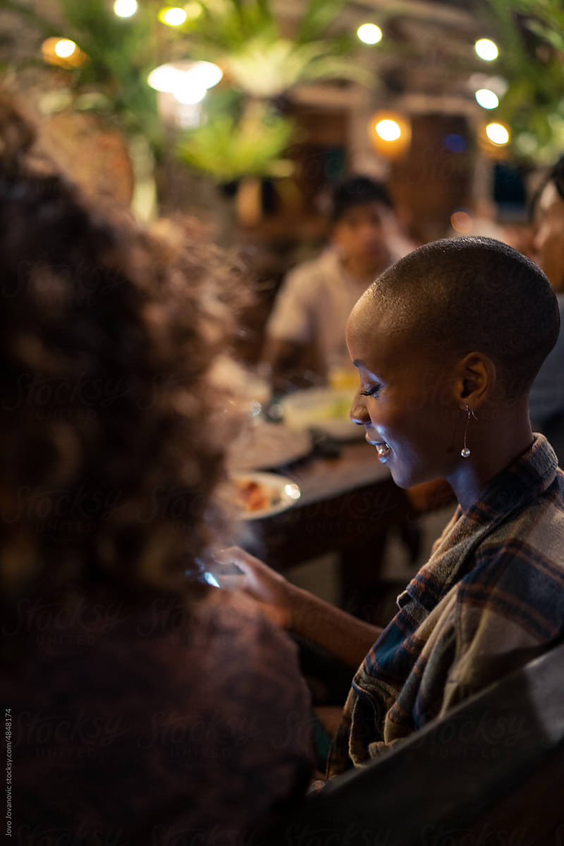 Black woman with shaved head using smartphone at dinner with friends