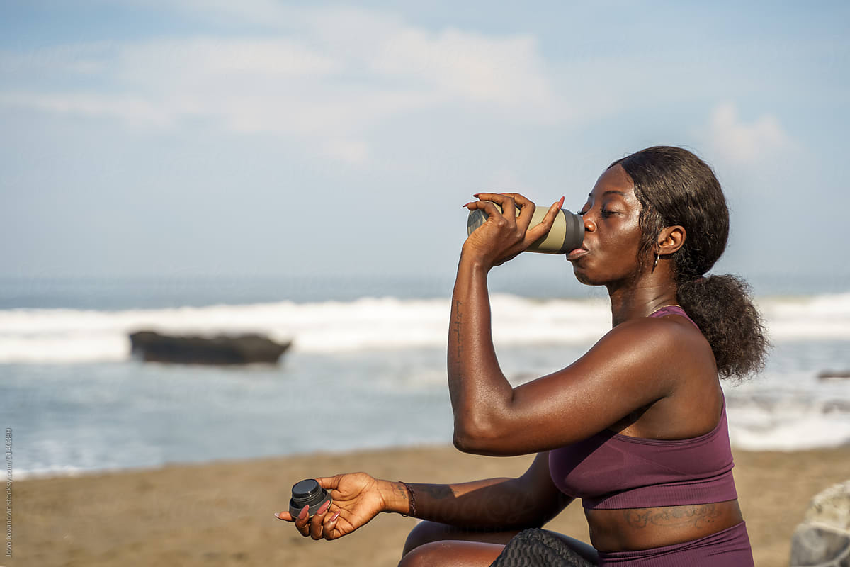 Woman drinking water from a flask sitting at the beach after workout