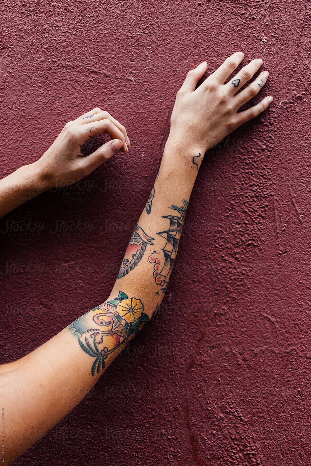 Tattooed arms reaching up a maroon concrete wall.