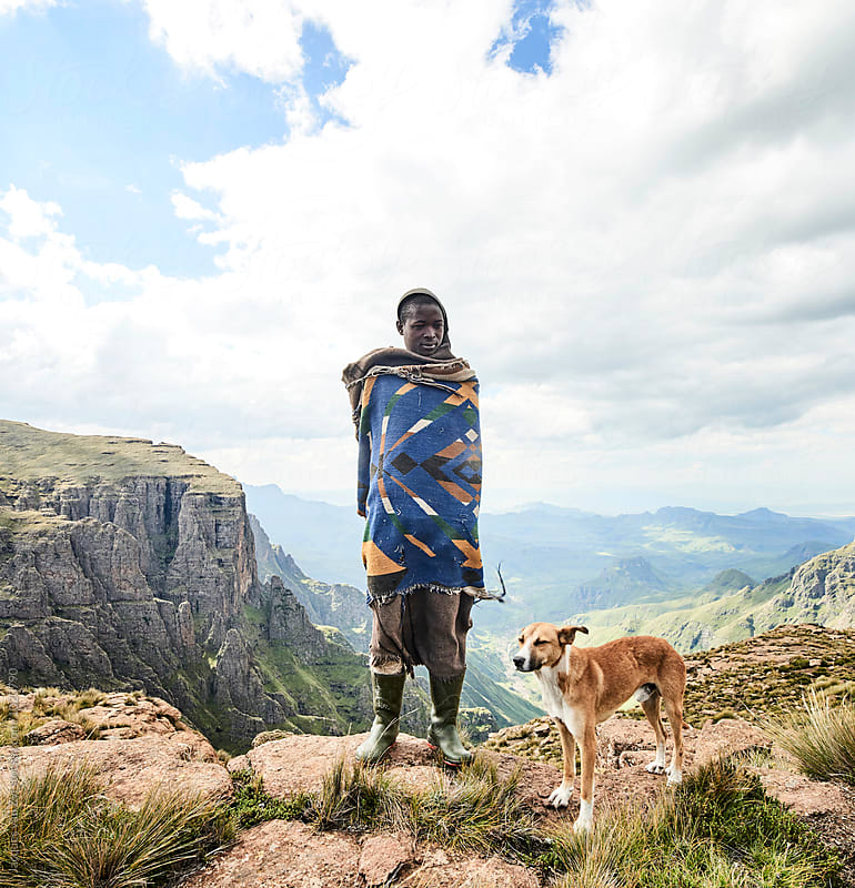 A Monsotho shepherd and clothed in traditional blanket, and walking stick, standing on a rock cliff edge with his dog in a beautiful mountain landscape.