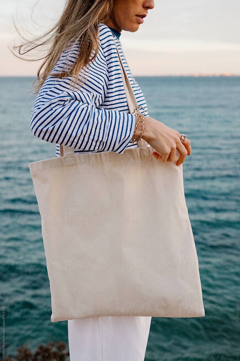 A young woman standing by the ocean, holding a plain white tote bag