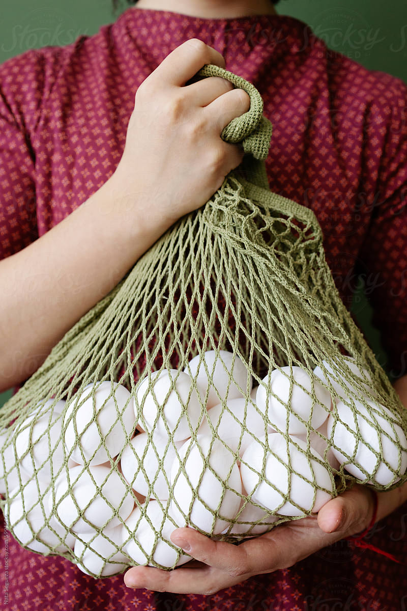 Person Holding a Mesh Bag Full of Eggs Against Green Background