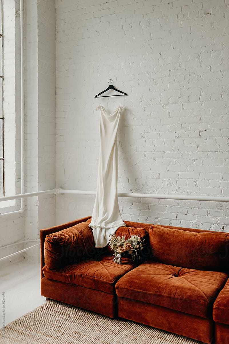 White wedding dress hanging on white brick wall of loft onto burnt orange velvet couch