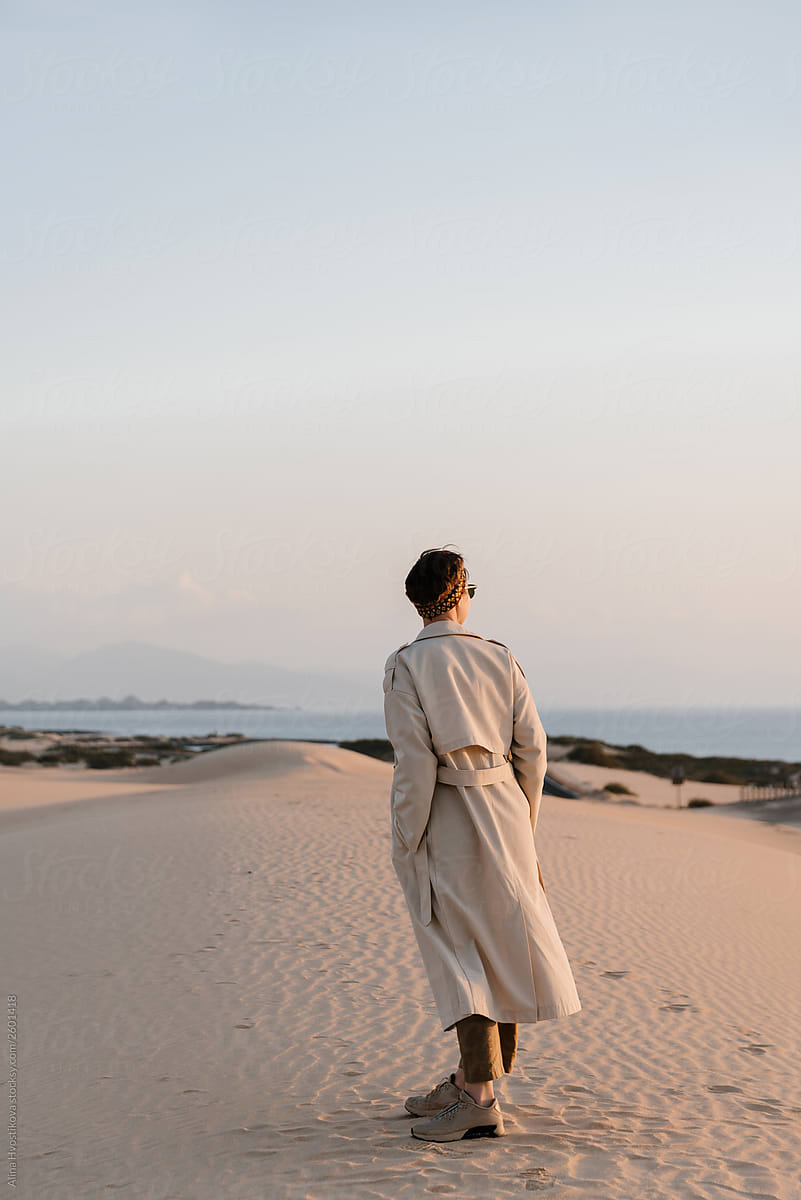 Brunette girl wearing beige trench coat walking among sand.