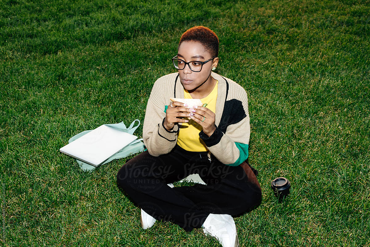 Female freelancer eating sandwich sitting on green lawn, direct flash