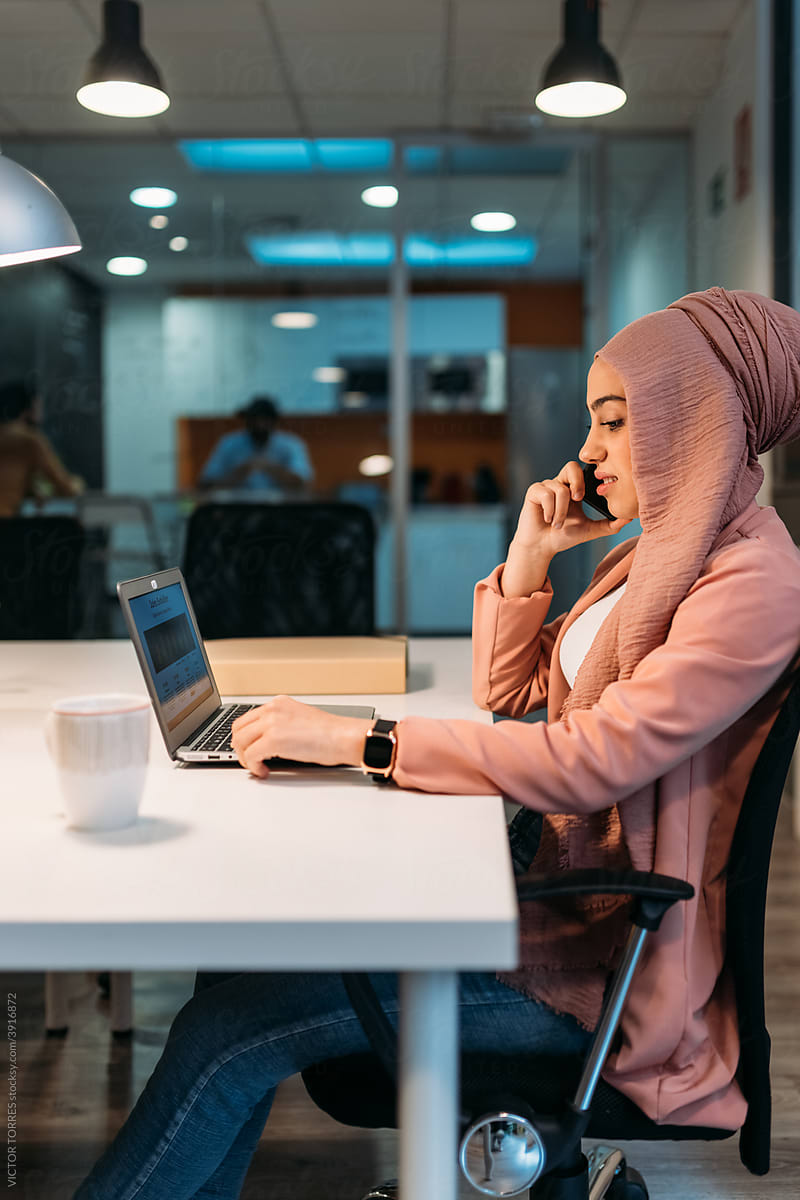 Smiling Muslim employee speaking on smartphone against laptop in office