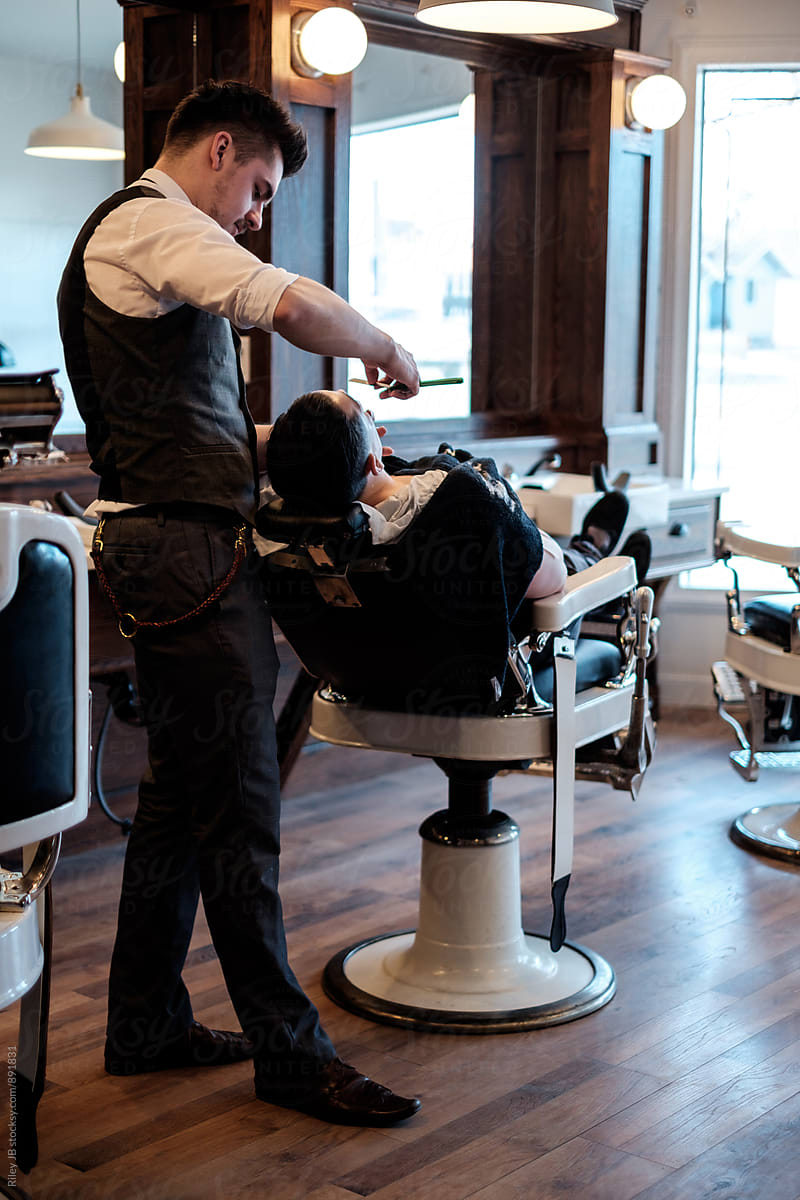 A gentleman barber shaving the face of a client that\'s reclined in a vintage barber chair.