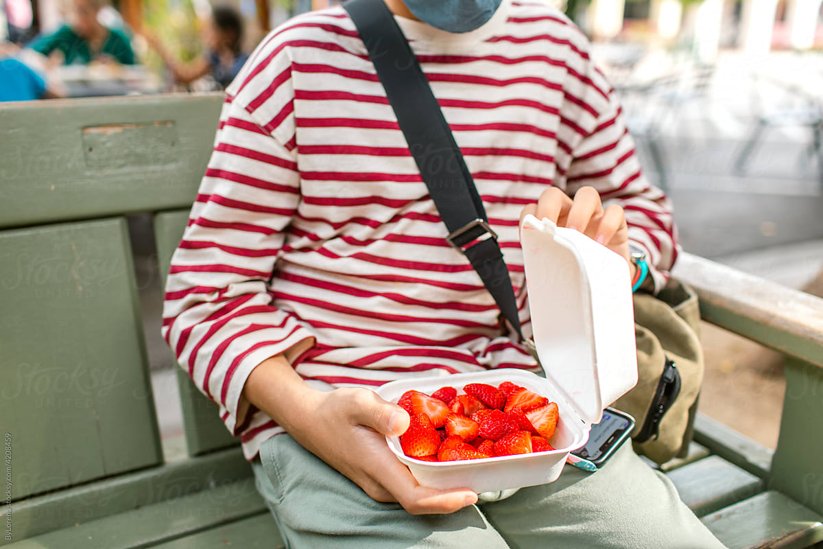 Cropped teen boy eating strawberries on bench in city