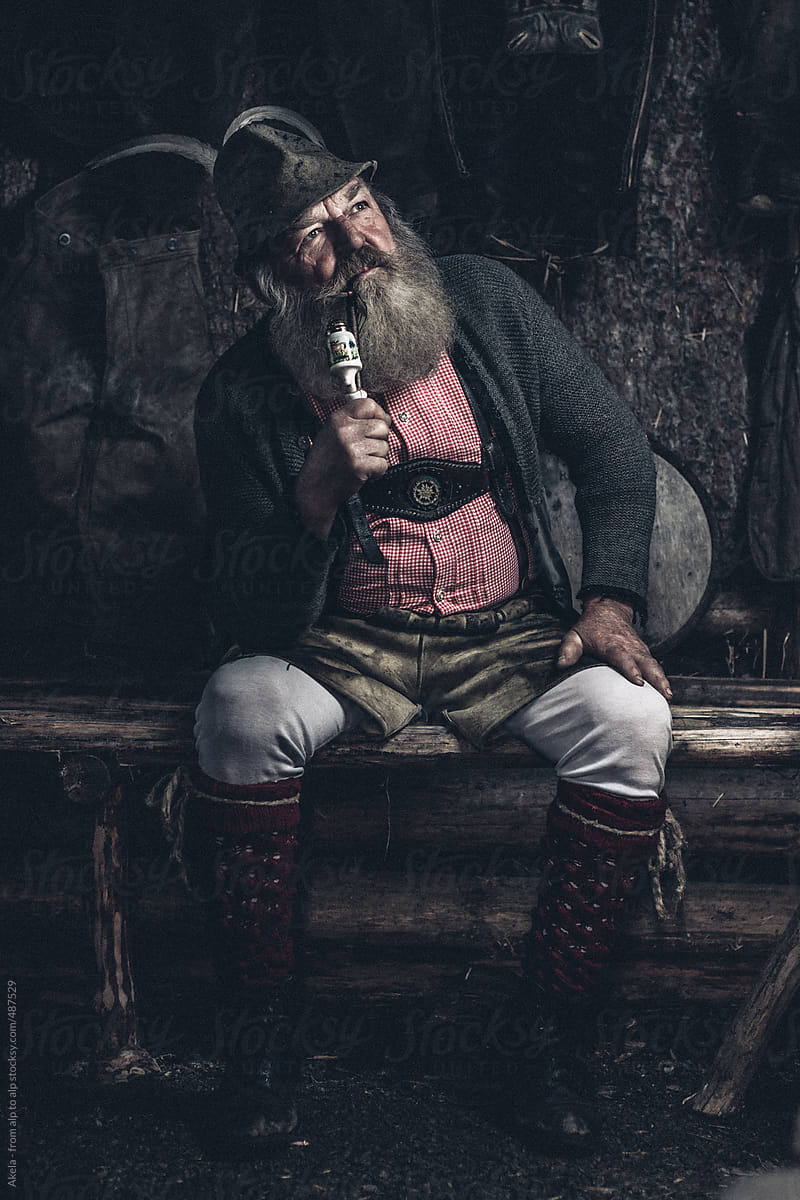old full bearded man in lederhosen sitting inside a wooden cabin smoking a pipe