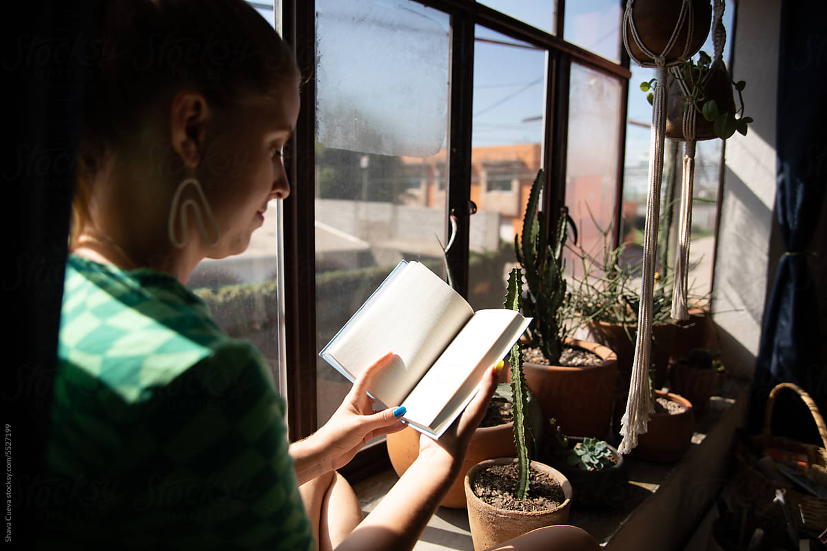 A woman reading a book next to a window with plants