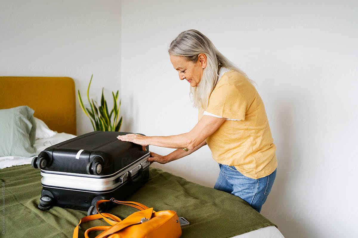 Gray-haired senior modern woman opening suitcase on bed