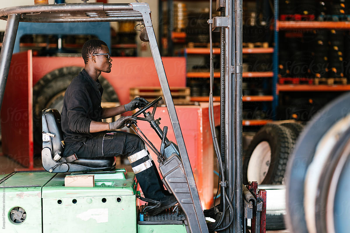 Black worker operating forklift truck at factory