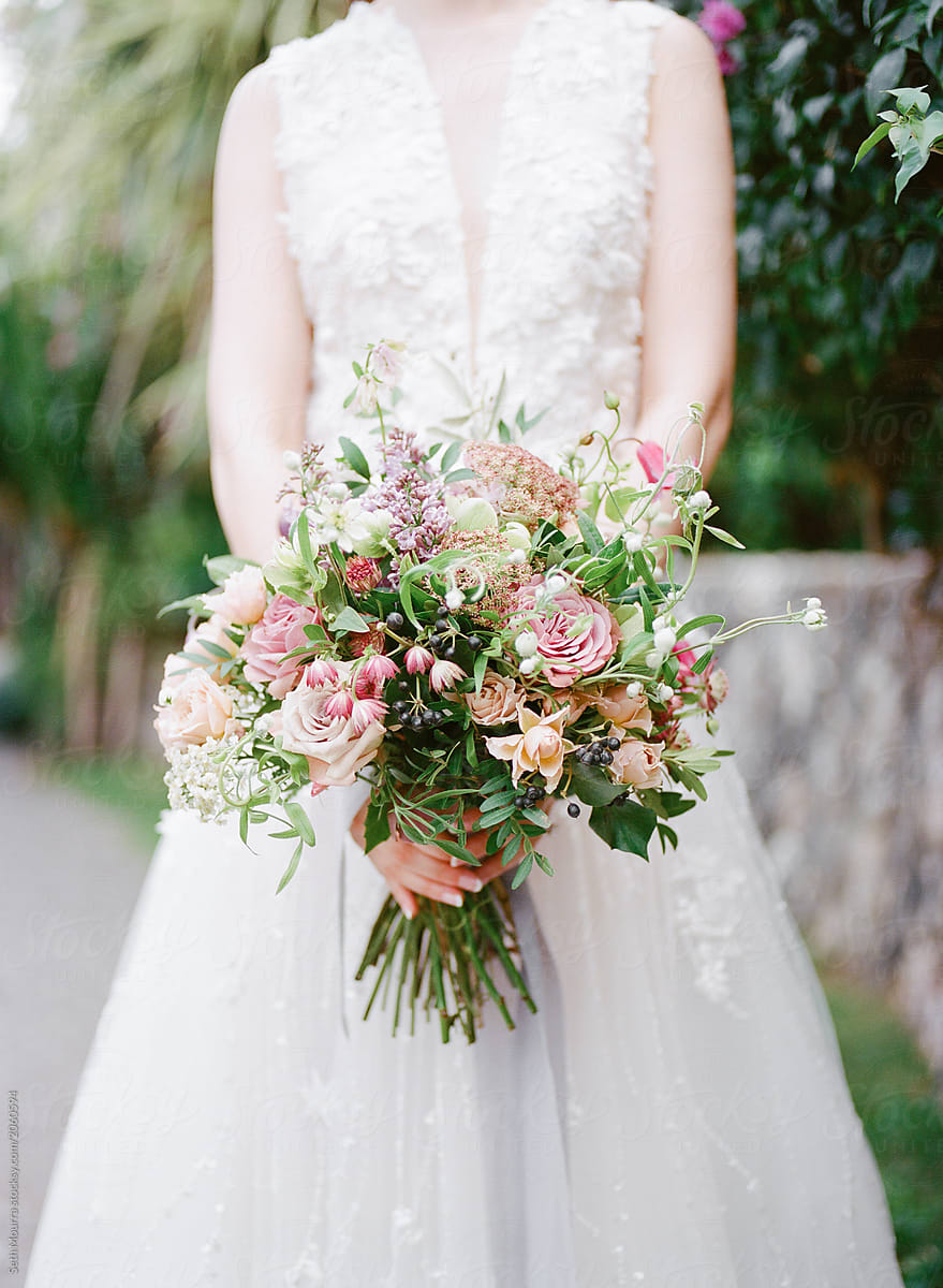A bride in a white detailed wedding gown holds a delicate green & pink floral bouquet