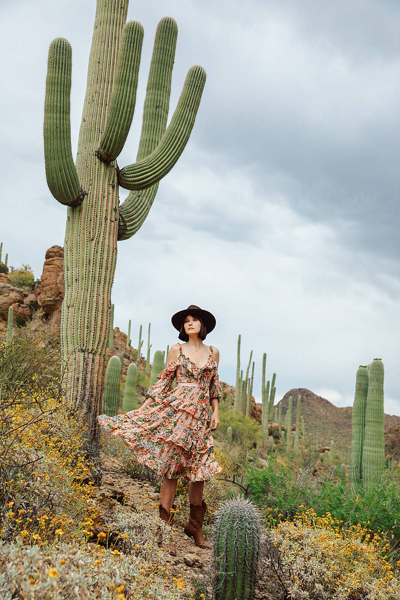 Young stylish cowgirl poses on rocks among saguaro cactus in the desert.