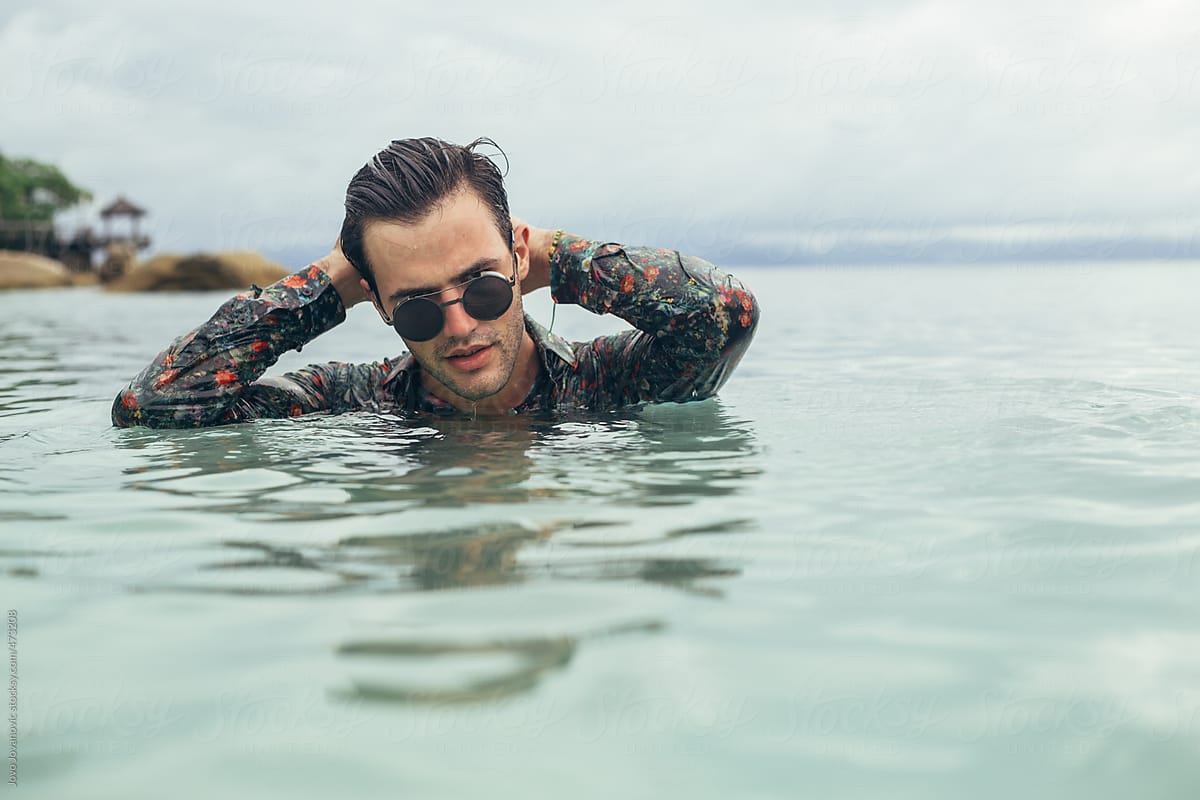 Wet young man fully clothed in ocean with sunglasses on slicking back his hair
