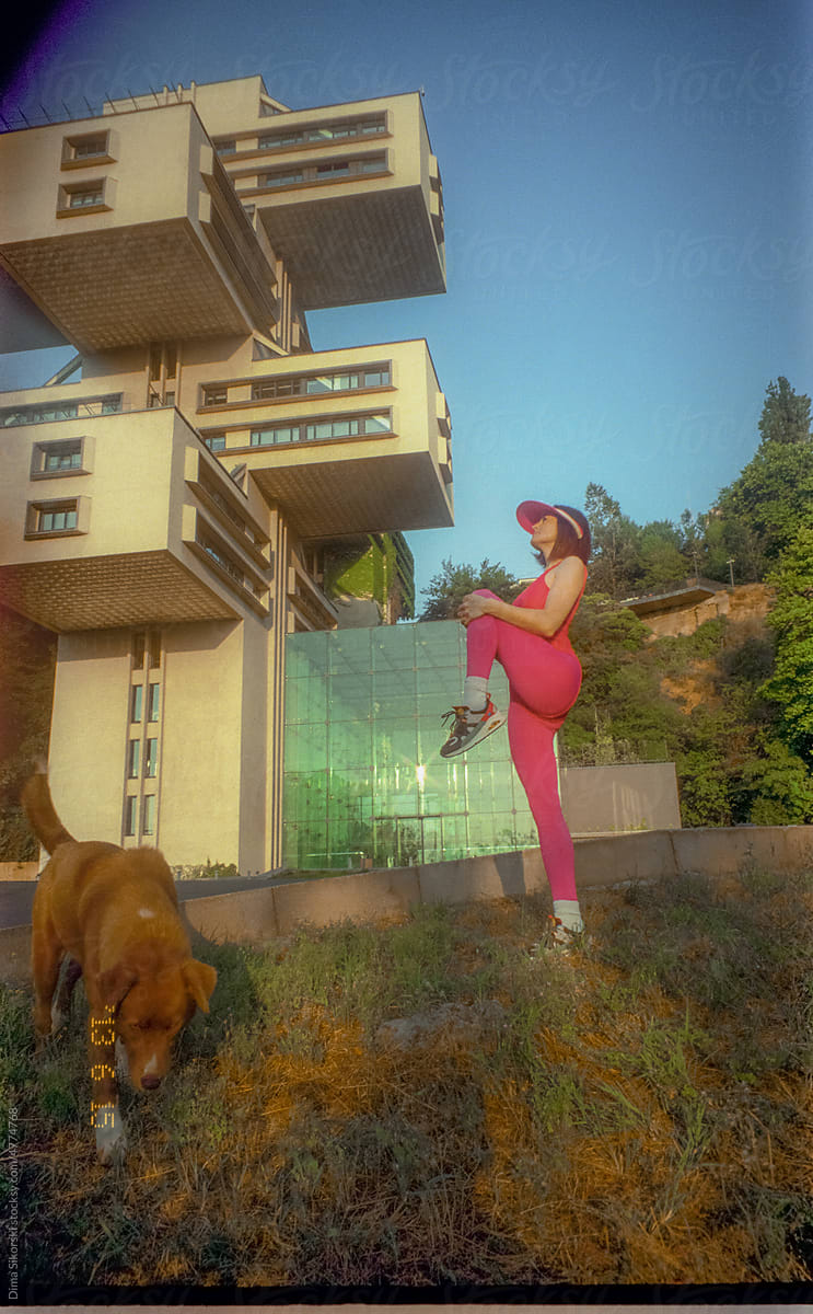 a girl does aerobics near a futuristic building with a dog