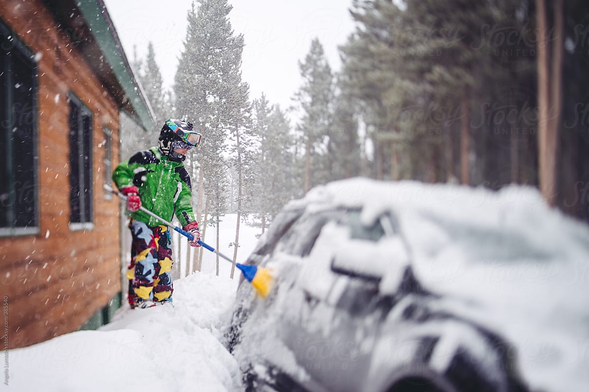 Boy in ski clothes sweeping snow off a car surrounded by deep snow