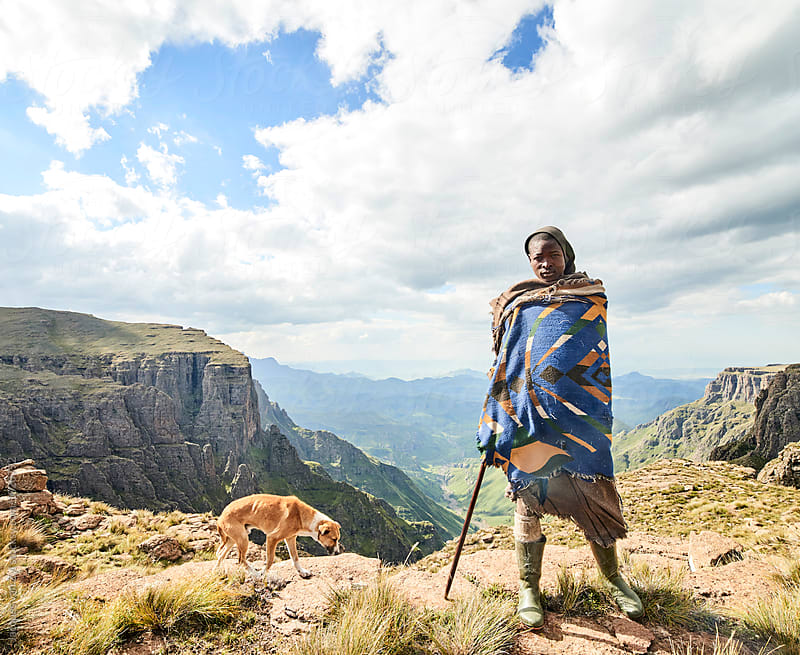 A Monsotho shepherd and clothed in traditional blanket, and walking stick, standing on a rock cliff edge with his dog in a beautiful mountain landscape.