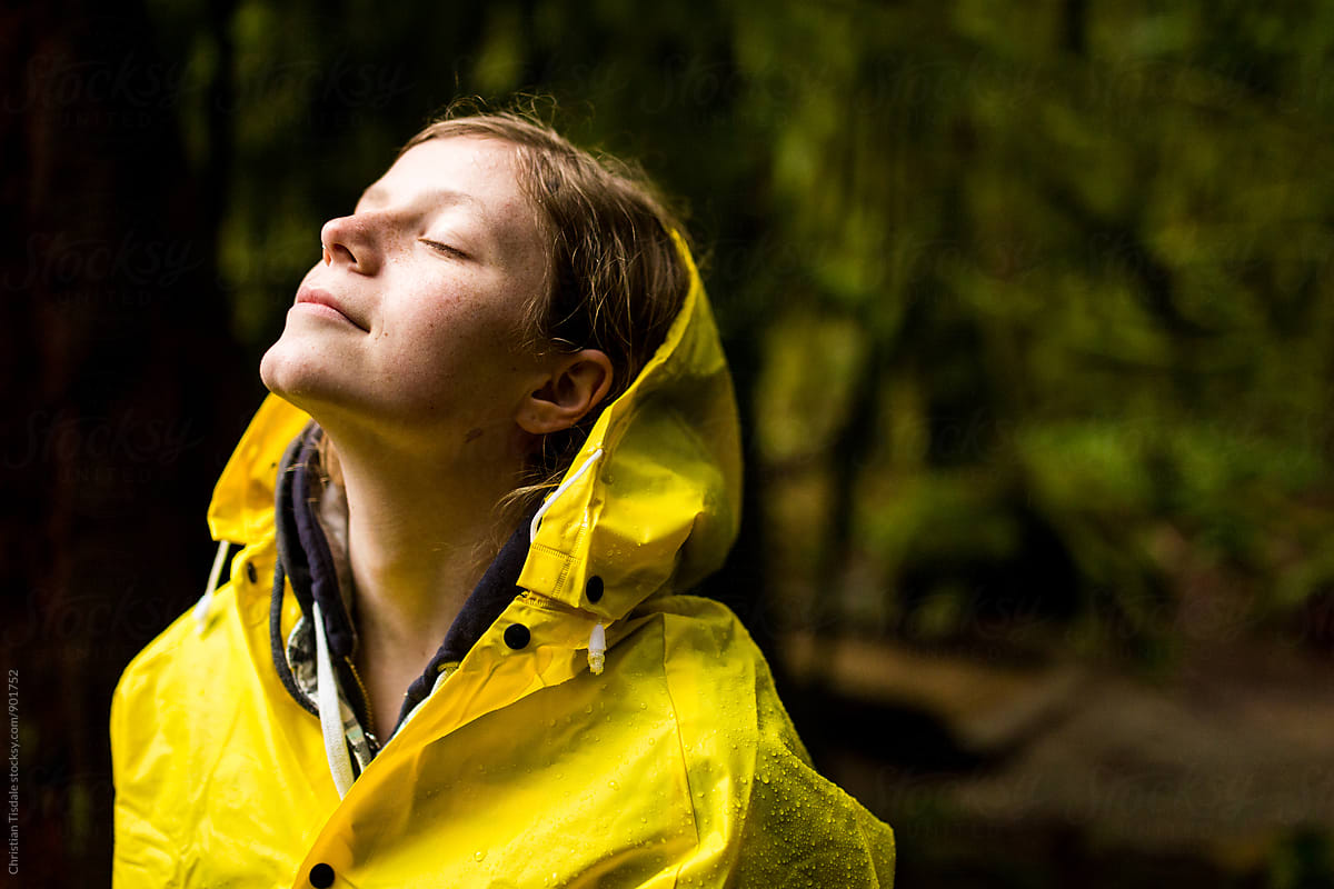 A young woman standing in a rainforest in a yellow raincoat with her head leaned back toward the sky
